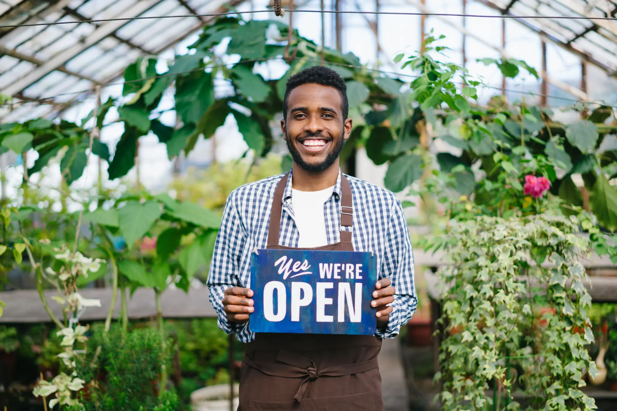 African American man is standing in greenhouse with we are open sign smiling looking at camera welcoming buyers for fresh flowers